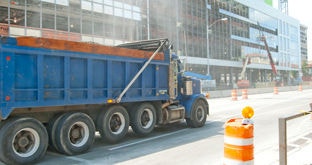 Construction dump truck on city street, traffic barrels, construction lifts and building under construction in the background